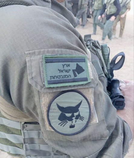 A terrorist Zionist pig soldier holding a patch with a map written on it, "The Jewish Promised Land," whose borders exte