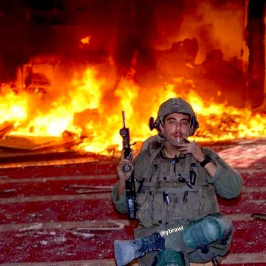 An Israeli soldier boasting about sharing a photo while sitting and jokingly smoking behind a mosque he set on fire in R