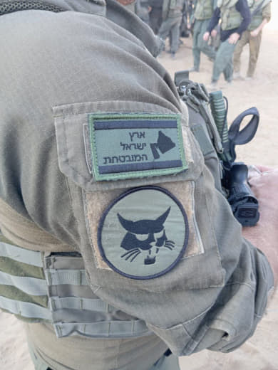 An Israeli occupation soldier is seen wearing a map logo labeled "Promised Land," with borders extending to Iraq, Saudi 